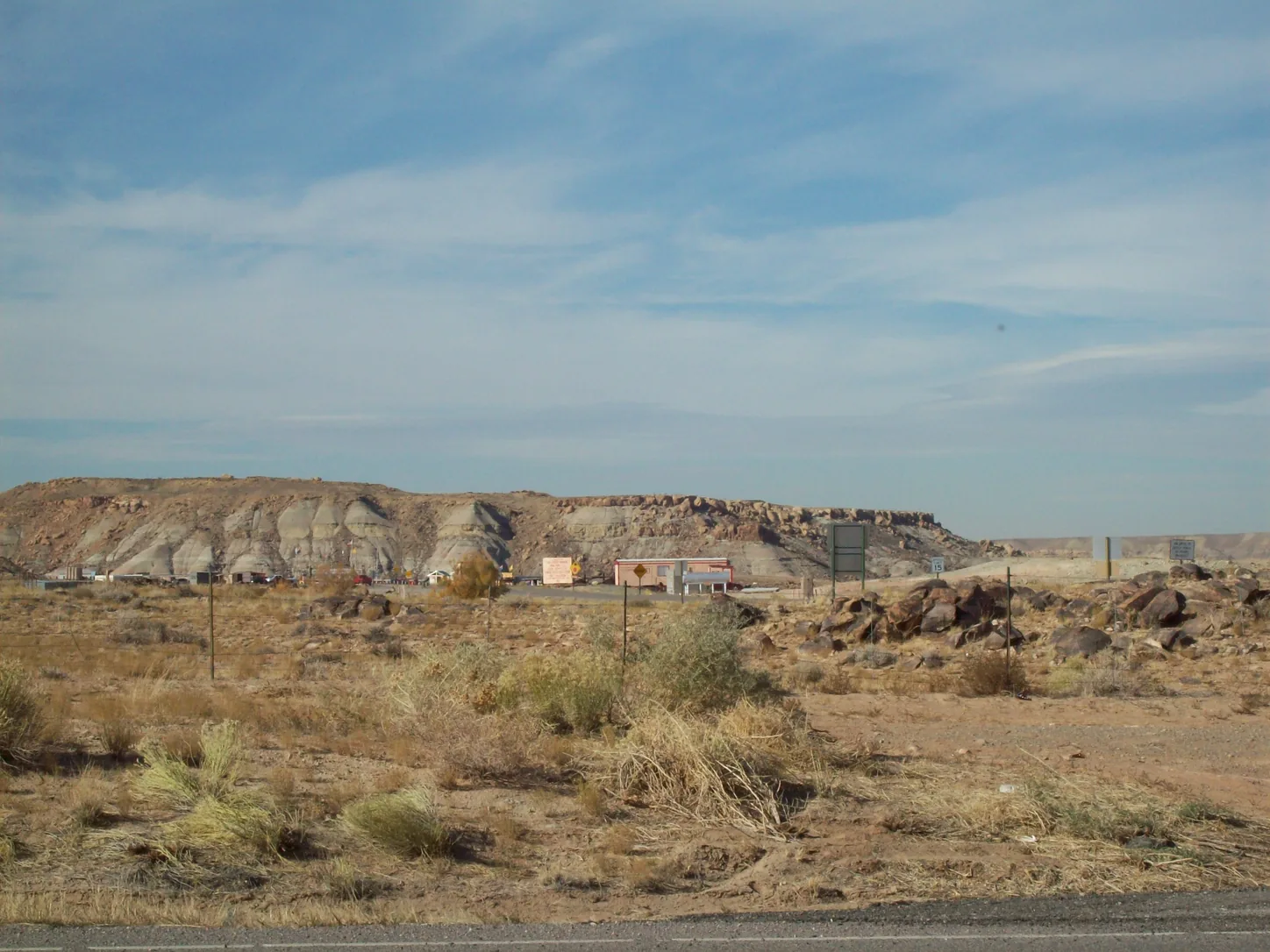 The view northwest from US-160 toward the monument: flat pale scrub and sage in the foreground, a low bluff behind, and the monument's buildings small and distant under a big open sky
