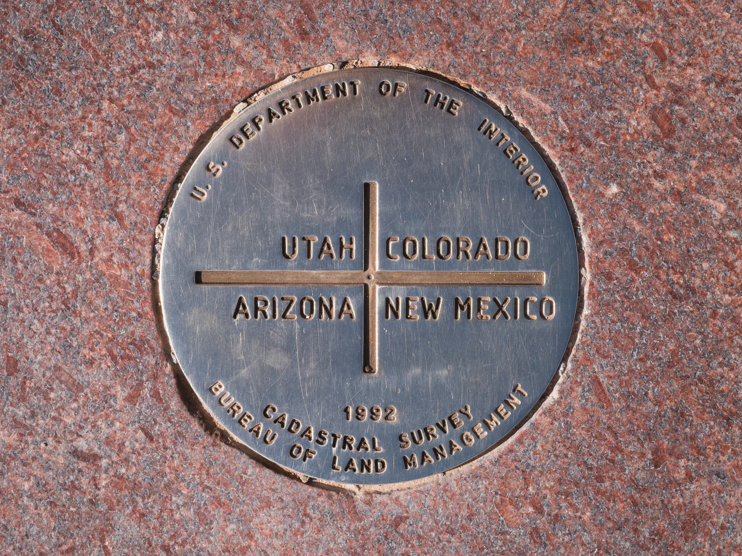 The survey disc at the centre of the monument: a bronze plate set flush into polished red granite, a raised cross dividing it into quadrants lettered UTAH, COLORADO, ARIZONA and NEW MEXICO, and around the foot of it 1992 CADASTRAL SURVEY, BUREAU OF LAND MANAGEMENT
