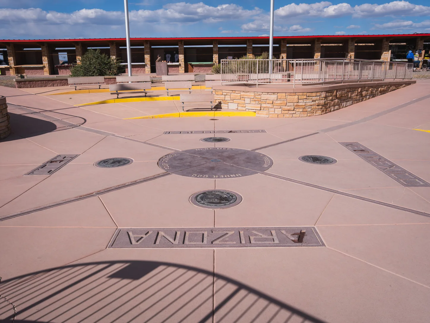 The flat paved plaza seen at ground level from the Arizona quadrant, ARIZONA cut into the paving in the foreground, bronze state seals set into each quadrant, and the long open craft galleries running along the far side